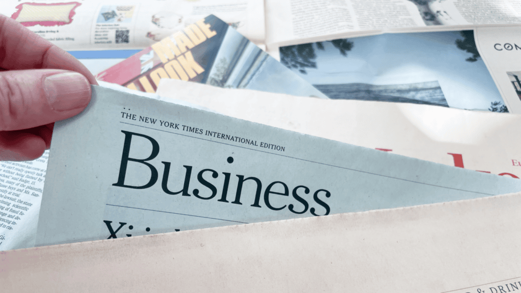 A hand holding open the Business section of The New York Times International Edition, surrounded by other newspapers and magazines.