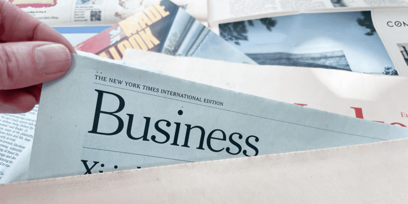 A hand holding open the Business section of The New York Times International Edition, surrounded by other newspapers and magazines.